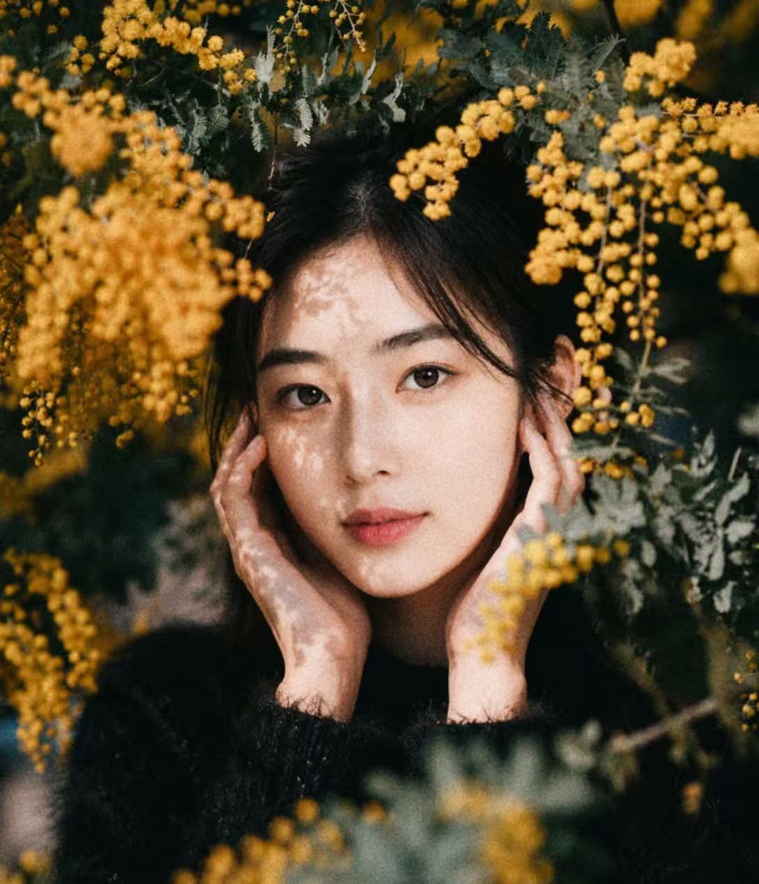 缇薇雅身穿黑色上衣，在金黄色花枝与绿叶间双手轻扶脸颊，望向镜头
