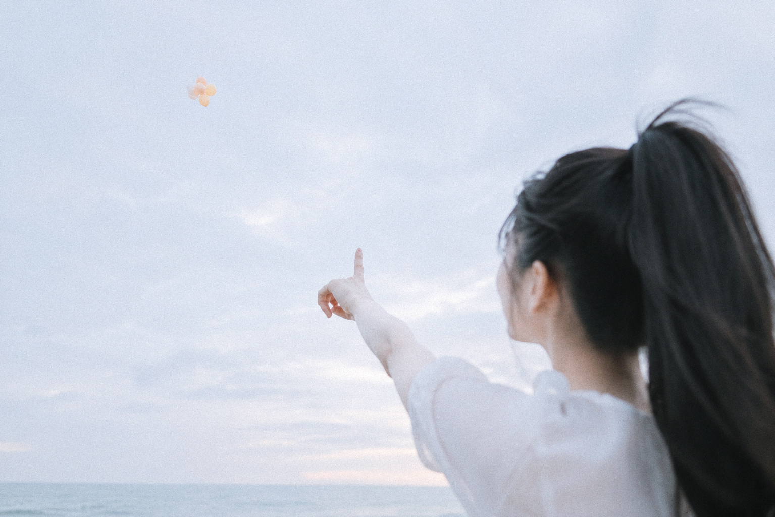 缇薇雅背对镜头望向天空，抬手指向远处的浅色气球